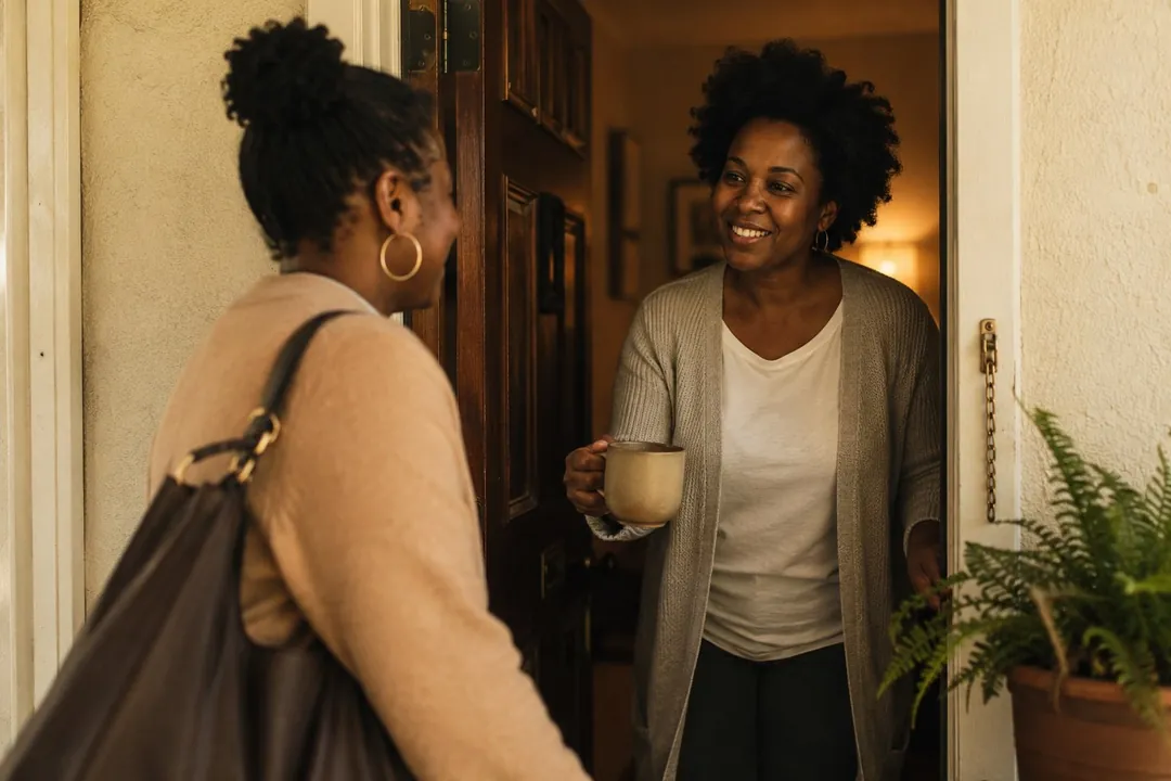 A woman with a warm smile welcoming a visitor at her front door, a cup of coffee in hand, in the golden evening light.